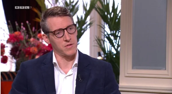 Een man met bril en colbert zit in een interieur met bloemen en planten. Links bovenin staat het logo van RTL 7.