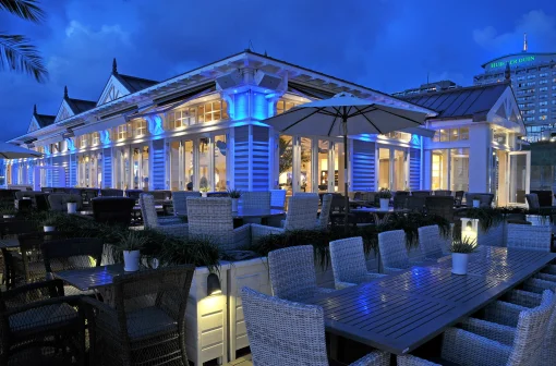Terras met rieten stoelen en tafels bij een verlicht restaurant in de schemering, met blauwe accenten en parasols.