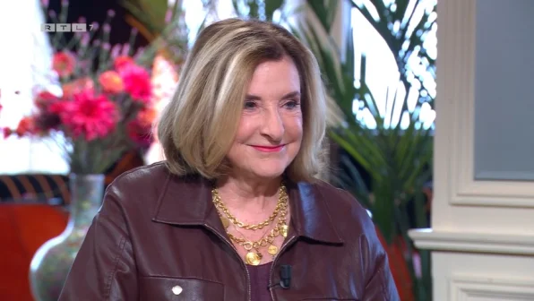 Een vrouw met blond haar draagt een bruine jas en gouden kettingen. Op de achtergrond zijn bloemen en planten zichtbaar.