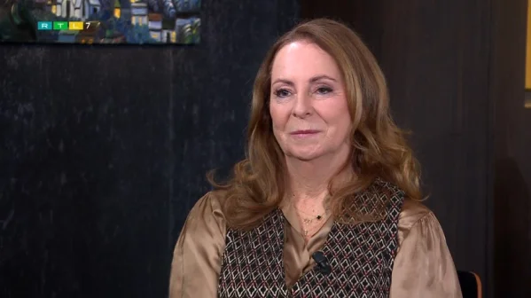 Een vrouw met lang bruin haar zit binnen, draagt een bruine blouse en vest. Op de achtergrond een schilderij en donkere muur.