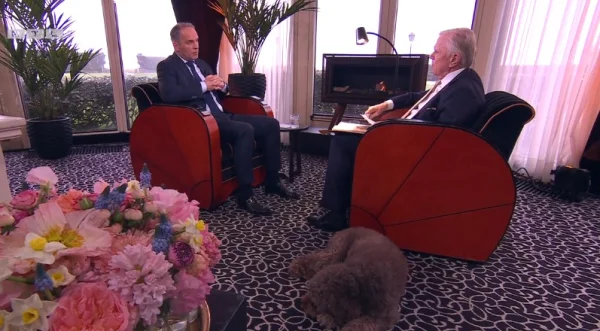 Twee mannen in gesprek in een luxe kamer met bloemen, een open haard en een slapende hond op de vloer.