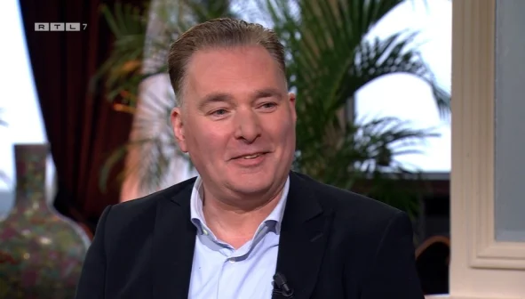 Een man in een pak lacht in een studio met planten op de achtergrond. Links bovenin staat het logo van RTL 7.