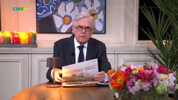 Een oudere man met bril leest een krant aan een tafel met bloemen en kaarsen. Achter hem hangt een kleurrijk schilderij.