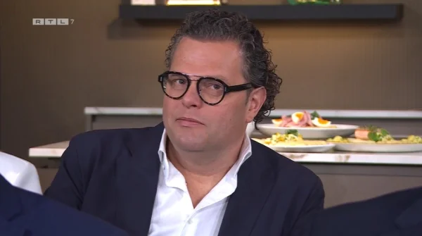 Een man met bril en krullend haar in een pak zit aan tafel. Op de achtergrond zijn borden met eten zichtbaar.