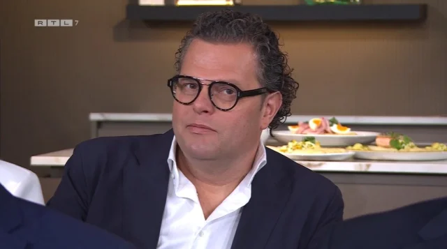 Een man met bril en krullend haar in een pak zit aan tafel. Op de achtergrond zijn borden met eten zichtbaar.
