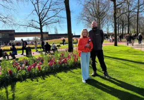 Twee mensen staan in een park met bloeiende tulpen en bomen. Op de achtergrond wandelen mensen en is een gebouw zichtbaar.