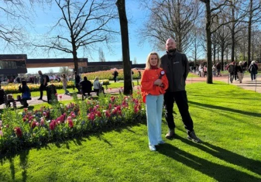 Twee mensen staan in een park met bloeiende tulpen en bomen. Op de achtergrond wandelen mensen en is een gebouw zichtbaar.