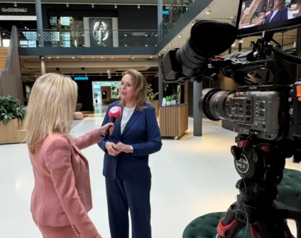 Twee vrouwen in een interview; een in roze pak met microfoon, de ander in blauw pak. Grote camera rechts, moderne kantooromgeving.