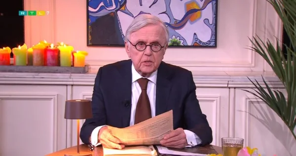 Een man in pak leest een krant aan tafel. Achter hem brandende kaarsen en een kleurrijk schilderij. Links een lamp, rechts een plant.