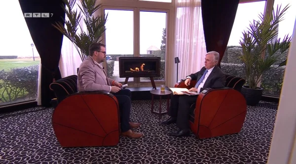 Twee mannen in gesprek in een luxe kamer met open haard, grote ramen en planten. Ze zitten in art-deco stoelen.