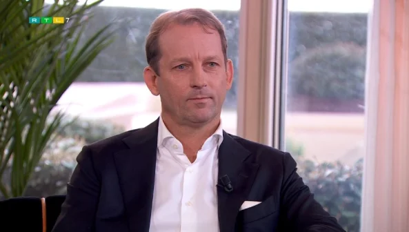 Een man in een pak zit voor een raam met planten op de achtergrond. Hij draagt een wit overhemd en een zwarte blazer.