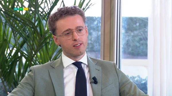 Een man in een pak met bril en microfoon, zittend voor een raam met planten op de achtergrond.