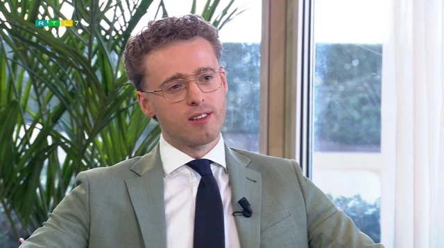 Een man in een pak met bril en microfoon, zittend voor een raam met planten op de achtergrond.