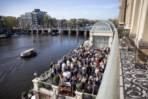 Een grote groep mensen verzameld op een terras aan het water, met een brug en gebouwen op de achtergrond.