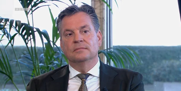 Een man in pak zit voor een raam met planten op de achtergrond. Hij kijkt serieus.
