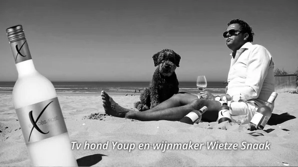 Man en hond zitten op strand met wijnflessen en glas. Grote fles rosé op voorgrond. Tekst onderaan beeld.