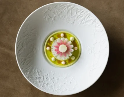 Wit bord met bloemvormige garnering in roze en wit, omringd door groene saus en kleine bolletjes.