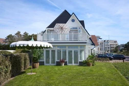 Wit huis met balkon, parasol en tuin met gras en planten. Auto's geparkeerd op de achtergrond onder een blauwe hemel.