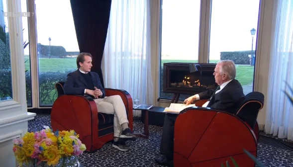 Twee mannen in gesprek in een luxe kamer met open haard, grote ramen en bloemen op tafel. Buiten is een groen gazon zichtbaar.