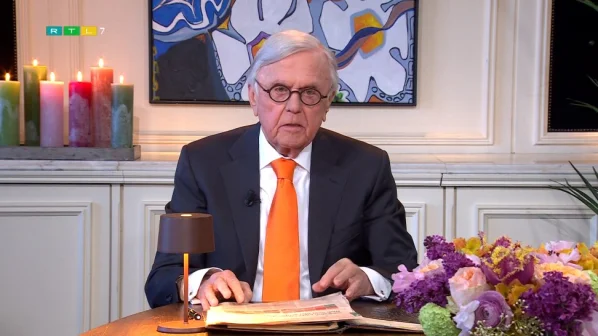 Een man in pak met oranje stropdas zit aan tafel met bloemen, kaarsen en een kunstwerk op de achtergrond.