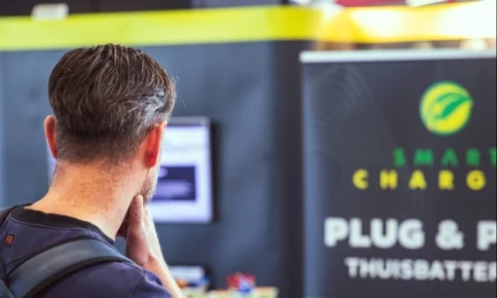 Een man met rugzak kijkt naar een bord met de tekst "Smart Charge" en "Plug & Play Thuisbatterij".