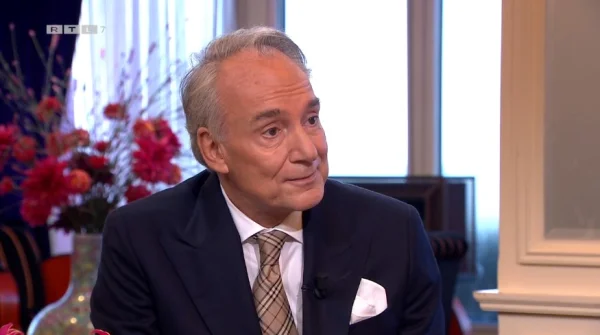 Een man in een donker pak met stropdas zit binnen, met bloemen op de achtergrond en een raam met lichtinval.