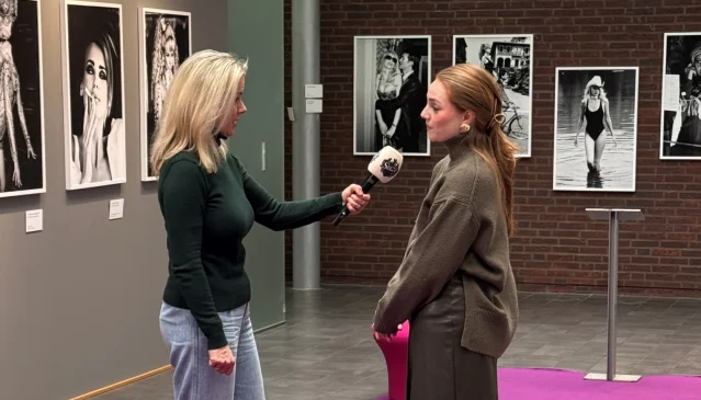 Twee vrouwen in een interview in een galerie met zwart-witfoto's aan de muur. Eén vrouw houdt een microfoon vast.