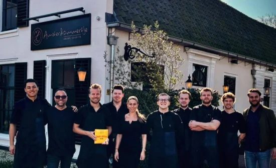 Een groep mensen poseert voor een restaurant met het bord "Amsterdammertje". Eén persoon houdt een gele gids vast.