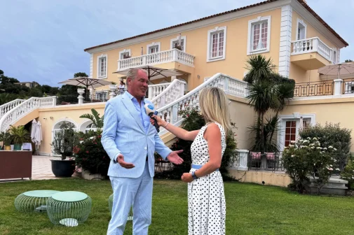 Twee mensen voor een grote villa; een vrouw in een witte jurk interviewt een man in een lichtblauw pak met een microfoon.
