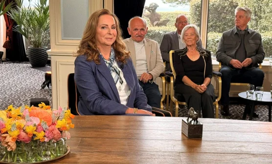 Een vrouw zit aan een tafel met bloemen en een beeldje. Vier mensen zitten op de achtergrond in een kamer met planten.