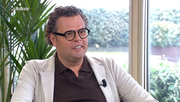 Een man met bril en microfoon zit binnen voor een raam met uitzicht op groen. Links een grote plant.