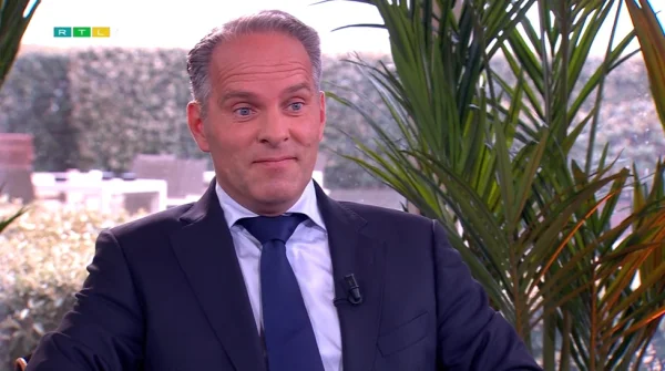 Een man in pak met een blauwe stropdas zit binnen, omringd door groene planten. Op de achtergrond is een tuin zichtbaar.