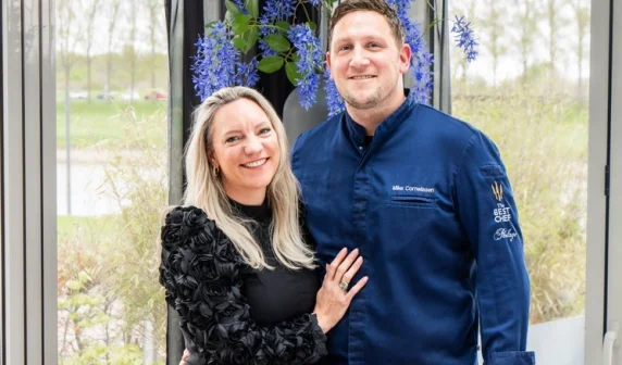 Een lachend stel poseert binnen bij een raam. De man draagt een blauwe chef-kok outfit, de vrouw een zwarte jurk.