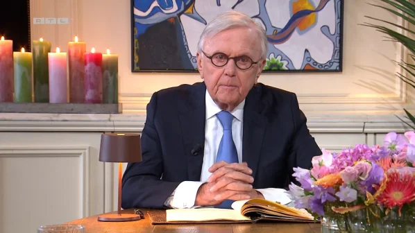 Een oudere man in pak zit aan tafel met een open boek. Op de achtergrond brandende kaarsen en een kleurrijk schilderij.
