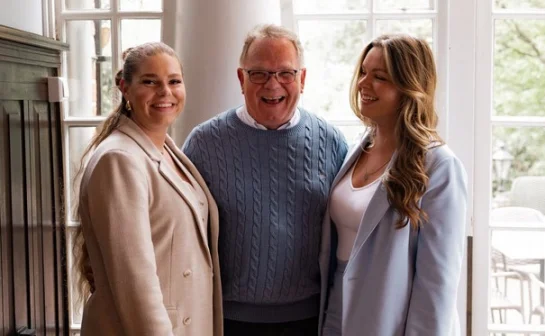 Een lachende man in een blauwe trui staat tussen twee vrouwen in beige en blauwe blazers, voor een raam met uitzicht op groen.