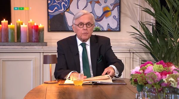 Een man in pak zit aan tafel met een boek. Op de achtergrond brandende kaarsen, een schilderij en bloemen op tafel.