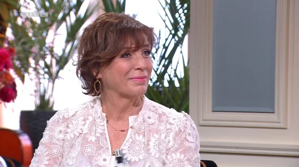 Een vrouw met kort bruin haar draagt een witte kanten blouse en zit binnen, met planten en een raam op de achtergrond.