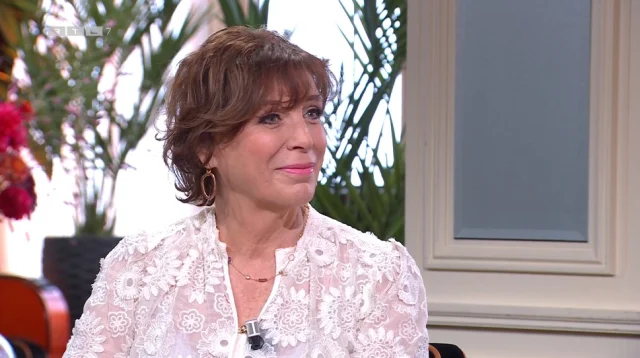 Een vrouw met kort bruin haar draagt een witte kanten blouse en zit binnen, met planten en een raam op de achtergrond.