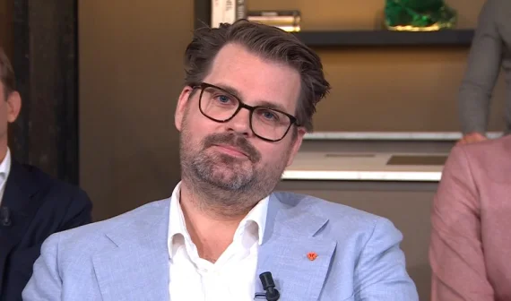 Een man met bril en baard in een lichtblauw pak zit in een studio. Op de achtergrond zijn vage figuren zichtbaar.
