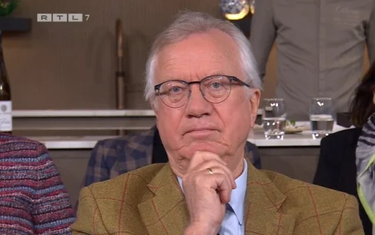 Een oudere man met bril en hand aan kin, zittend in een studio. Op de achtergrond glazen en een persoon in een grijs overhemd.