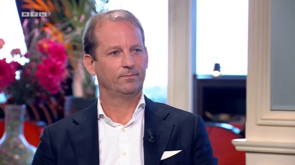 Een man in een pak zit in een studio met kleurrijke bloemen op de achtergrond. Links boven is een RTL7-logo zichtbaar.