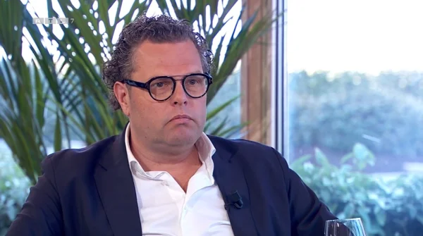 Een man met bril en krullend haar in een pak zit voor een raam met planten op de achtergrond.