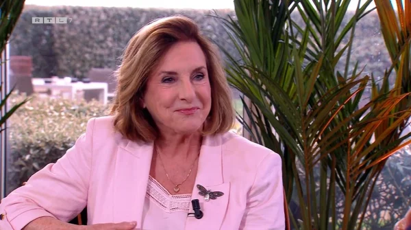 Een vrouw in een roze blazer zit binnen naast groene planten, glimlachend. Op de achtergrond is een heg zichtbaar.