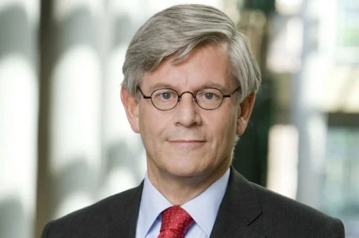 Een man met grijs haar en bril draagt een donker pak en rode stropdas. Hij staat voor een wazige achtergrond met lichtinval.