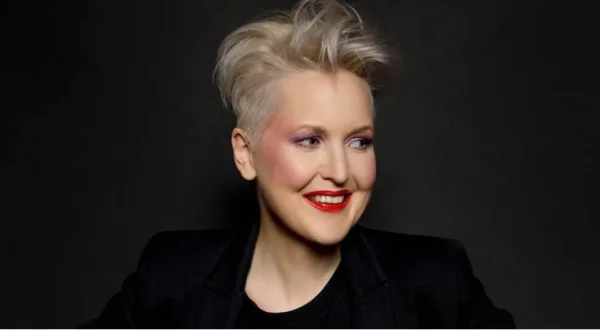 Een persoon met kort blond haar en rode lippenstift glimlacht, gekleed in een zwart jasje tegen een donkere achtergrond.