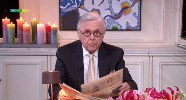 Een oudere man in pak leest een krant aan tafel. Op de achtergrond brandende kaarsen en een kleurrijk schilderij.