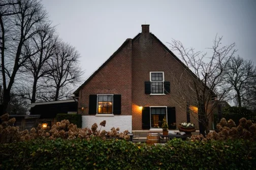 Bakstenen huis met donkere luiken, omgeven door kale bomen en struiken. Buitenverlichting aan, met een kerstboom op het terras.