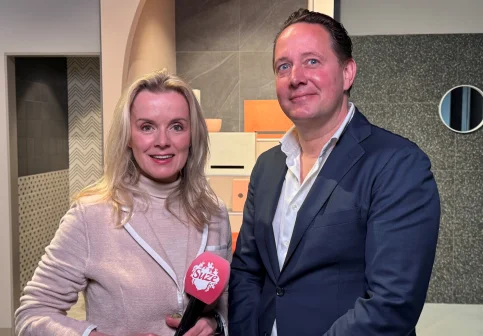 Twee mensen poseren binnen; vrouw met microfoon, man in pak. Achtergrond met tegels in verschillende patronen en kleuren.