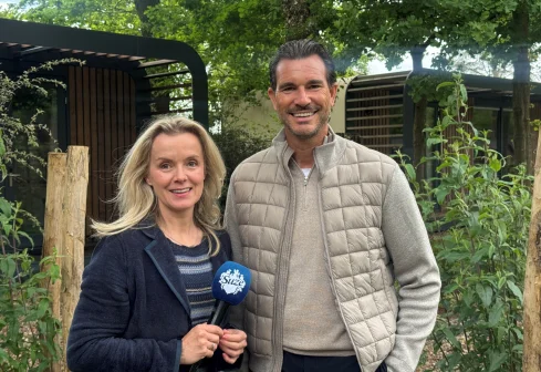 Twee mensen poseren buiten met een microfoon, omringd door groen en houten structuren. Ze lachen naar de camera.