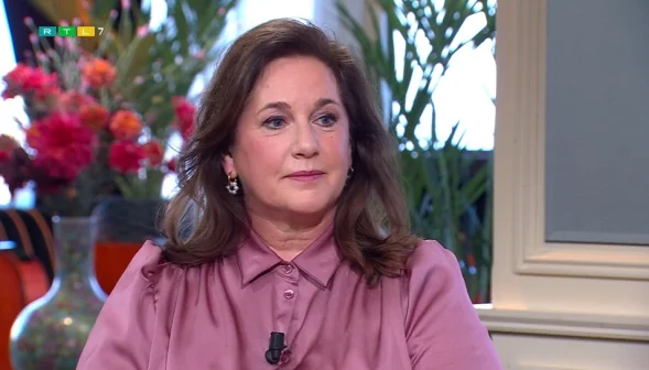 Een vrouw in een roze blouse zit in een studio met bloemen op de achtergrond. Ze kijkt naar links.